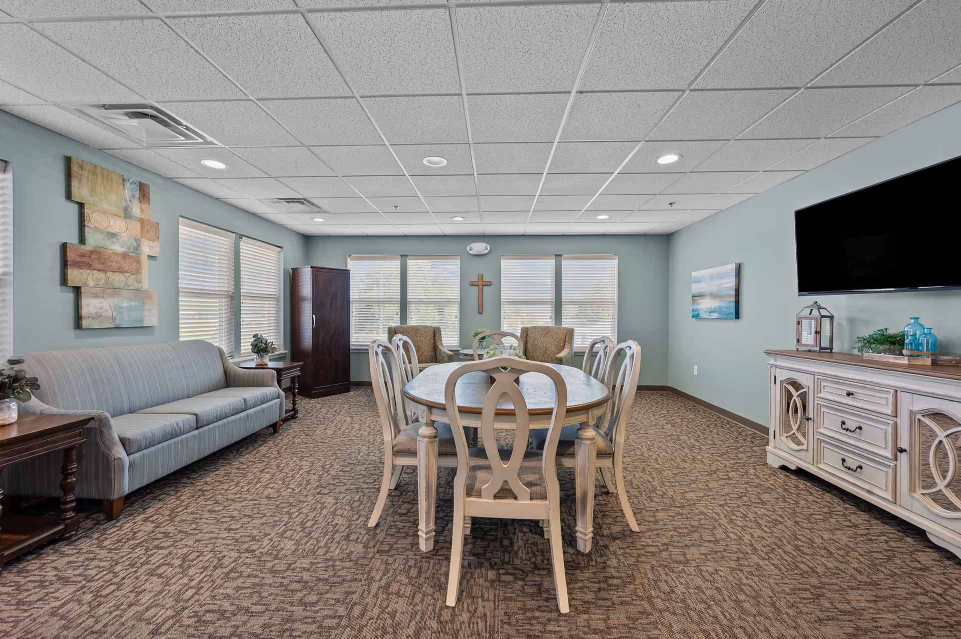 A senior living facility common room with a light blue sofa on the left, a wooden dining table with six chairs in the center, two armchairs near the windows at the back, and a white sideboard with decorative items and a flat-screen TV mounted above it on the right. The walls are painted light green, and there is a cross hanging on the wall between the windows.