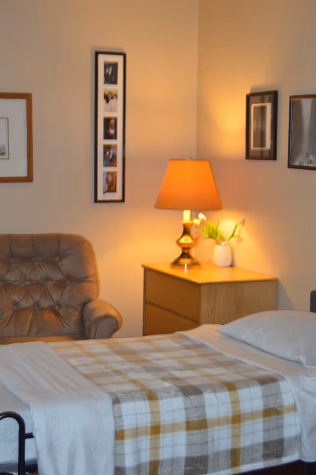 A cozy bedroom with a single bed covered with a plaid blanket in shades of beige, white, and yellow. Next to the bed is a wooden nightstand with a lit table lamp and a small white vase with flowers. To the left of the nightstand is a cushioned armchair. The walls are decorated with framed pictures.