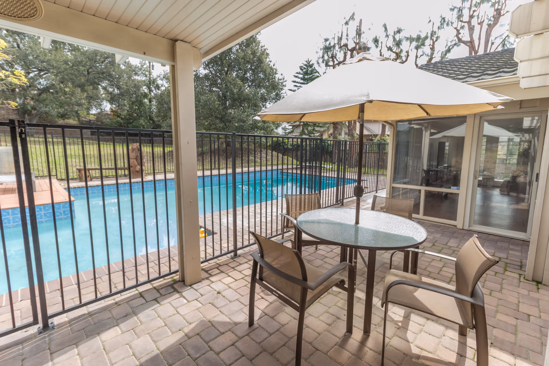 Covered patio with a round glass table, umbrella and chairs overlooking a fenced swimming pool and lawn.