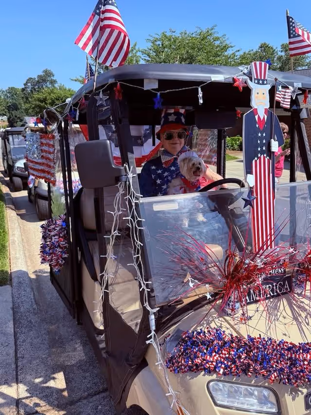 A decorated golf cart with American flags and patriotic decorations driven by a person in patriotic clothing holding a small dog during an outdoor parade.