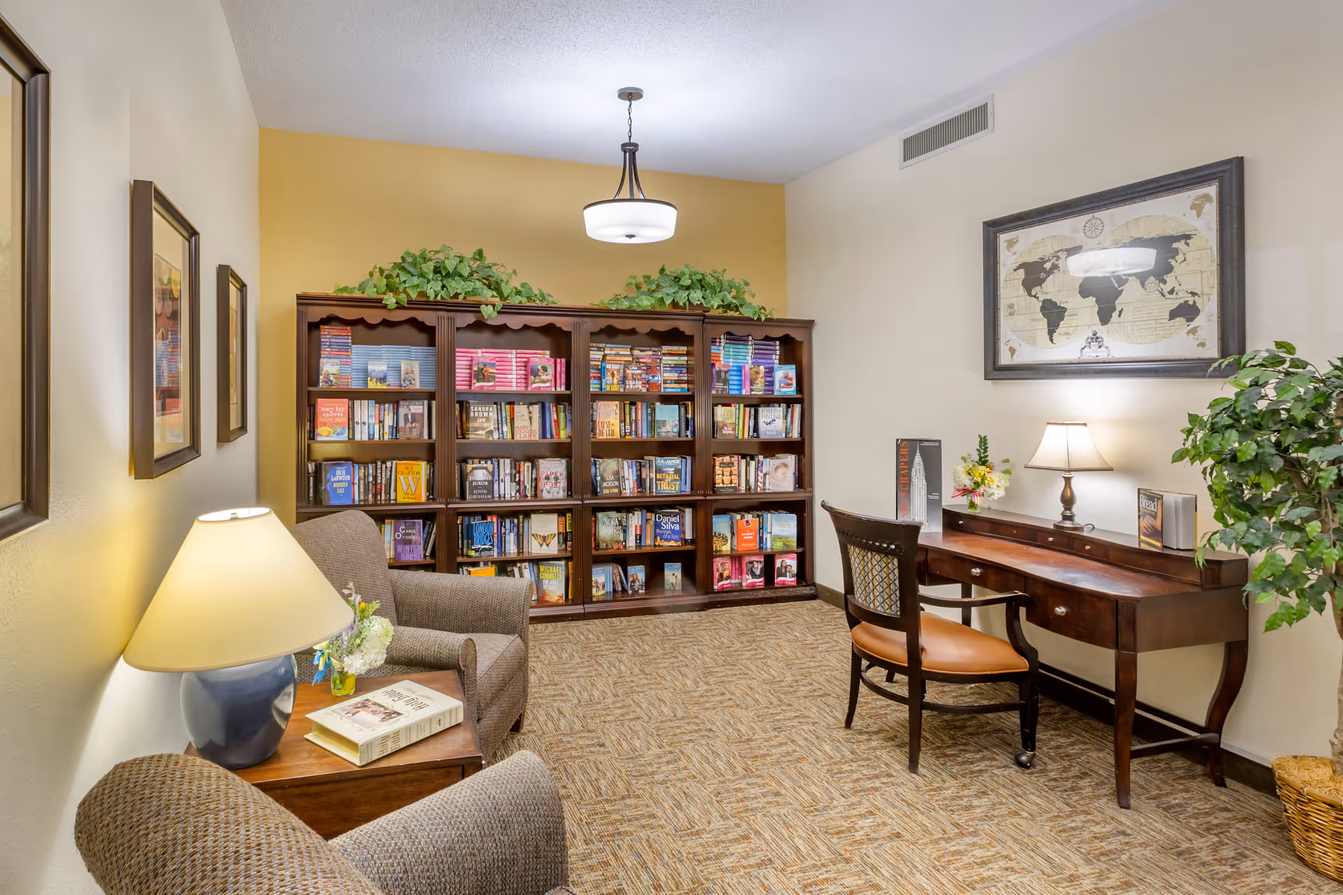 Cozy interior reading room with bookshelves, armchairs, a desk with a lamp, and framed artwork.