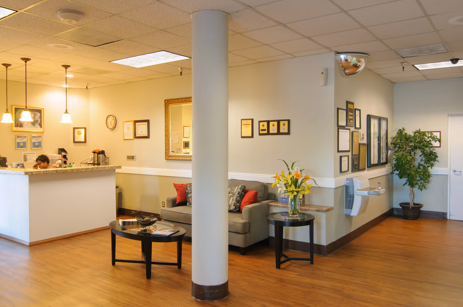 Well-lit reception and seating area with a front desk, sofa, coffee tables, vase of flowers, and framed certificates on the walls.