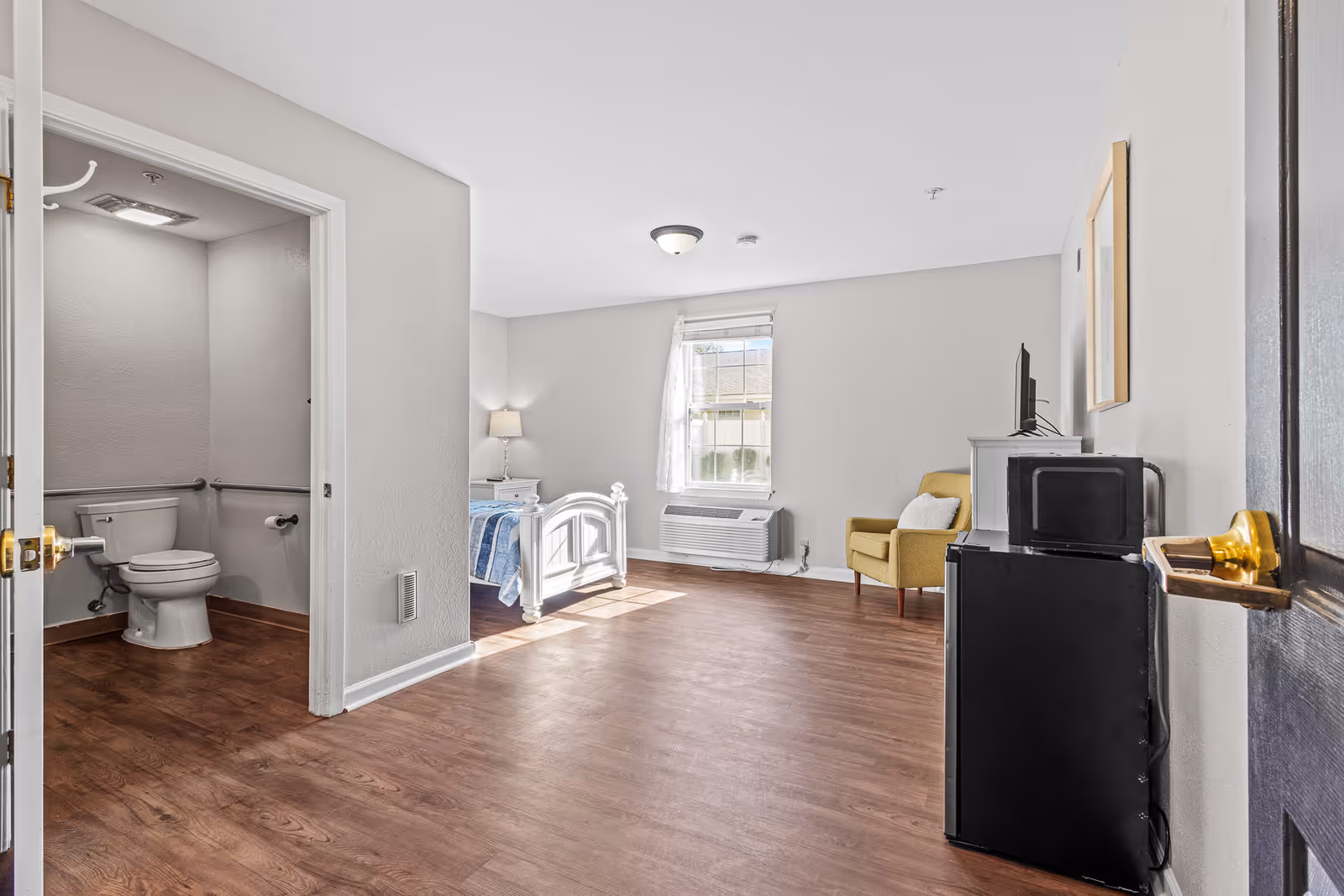 Bright studio bedroom with a single bed, yellow armchair, TV and an open accessible bathroom.