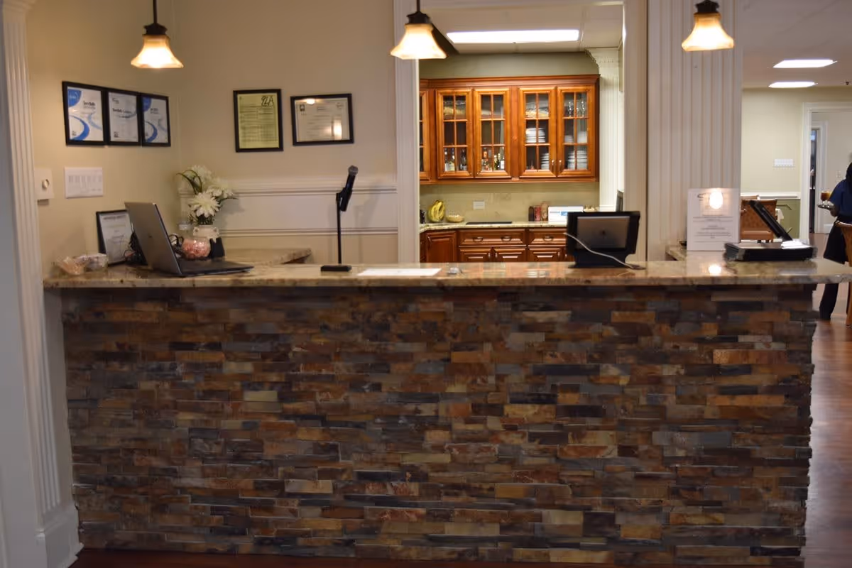 Reception desk area with a stone facade and granite countertop, featuring a laptop, a microphone, and some documents. Behind the desk is a kitchen area with wooden cabinets and glass doors displaying dishes. The walls have framed certificates and a vase with white flowers. Two hanging pendant lights illuminate the desk area.