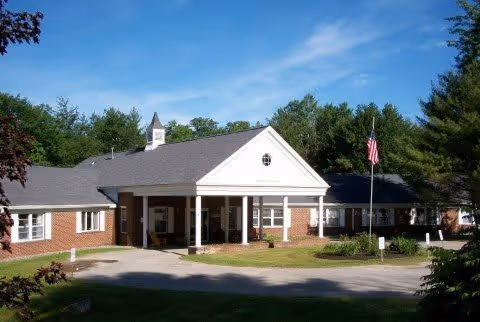 Exterior view of a single-story brick building with a white portico entrance, surrounded by trees and greenery under a clear blue sky. An American flag is visible on a flagpole near the entrance.