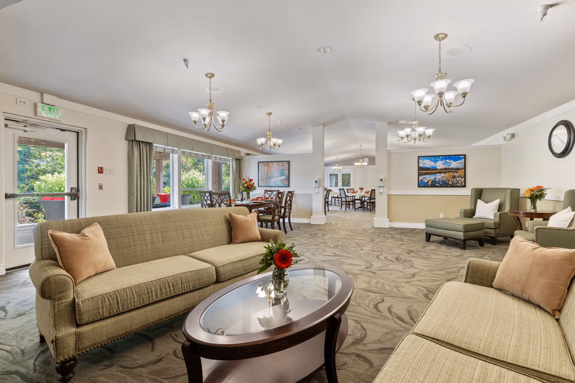A spacious and well-lit common area in a senior living facility featuring two beige sofas with brown pillows facing a glass-top coffee table with a vase of red flowers. In the background, there are dining tables with chairs, floral arrangements, and large windows letting in natural light. The room has beige walls, carpeted floors with a subtle pattern, multiple chandeliers, and framed landscape artwork on the walls.