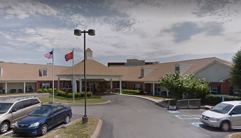 Exterior view of a single-story senior living facility building with a covered entrance, two flagpoles displaying the American and Tennessee state flags, several parked cars, and a partly cloudy sky.