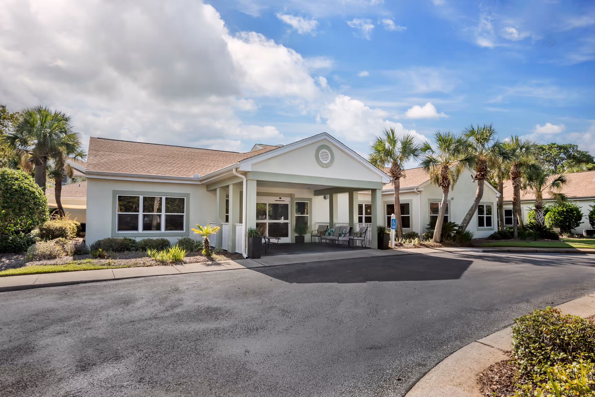 Exterior view of a single-story building with a covered entrance, surrounded by palm trees and landscaping under a partly cloudy sky.