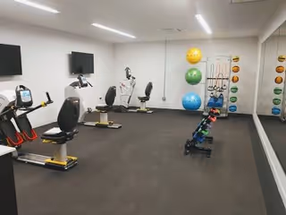 A fitness room with exercise equipment including stationary bikes, a rack of colorful dumbbells, large exercise balls in yellow, green, and blue, and a wall-mounted television. The room has a large mirror on one wall and bright overhead lighting.