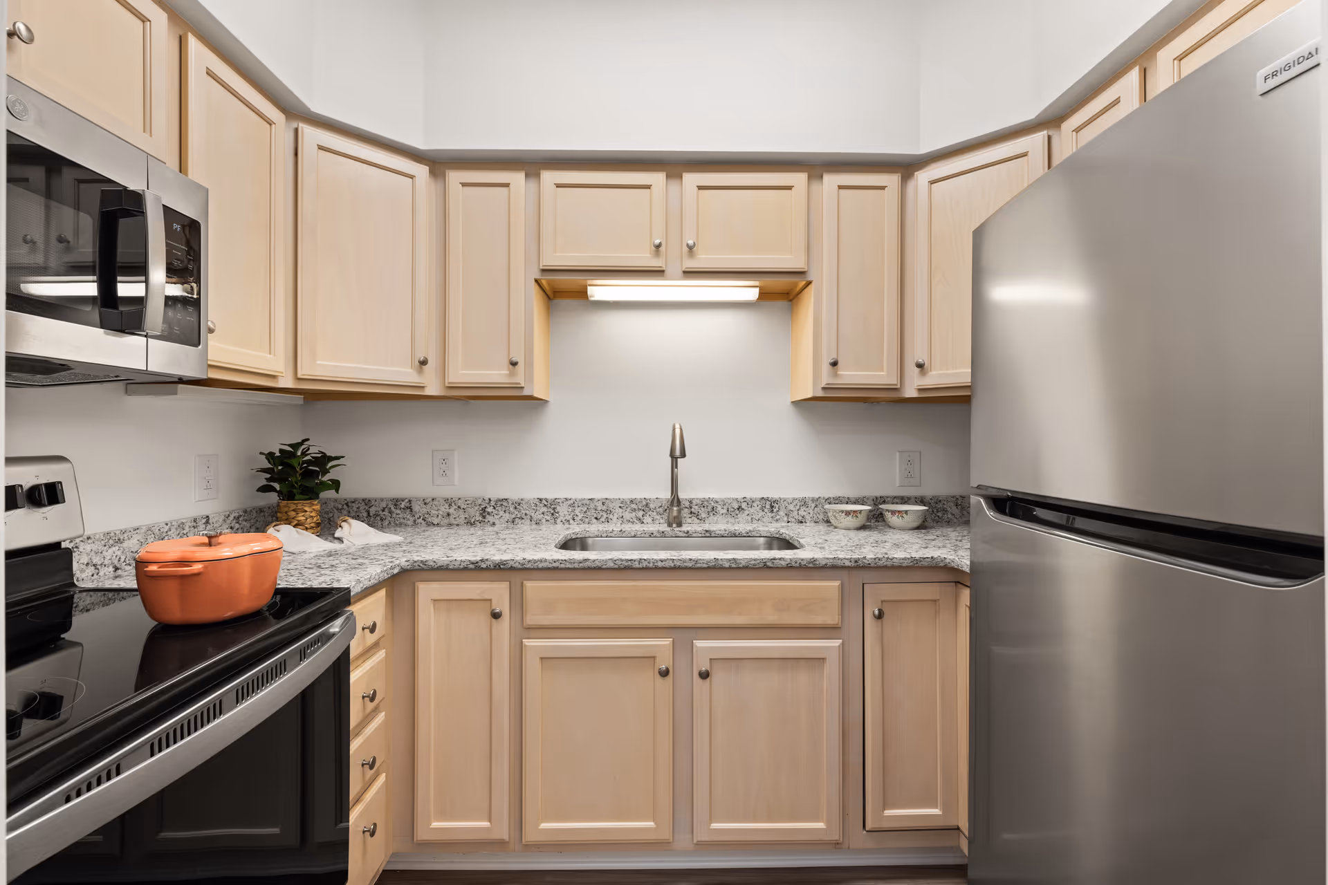 A modern kitchen with light wood cabinets, granite countertops, a stainless steel refrigerator, microwave, and stove. There is a sink centered under a cabinet with a light, a small potted plant, and two bowls on the countertop.