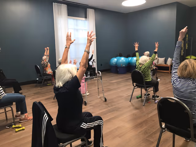 A group of elderly individuals seated on chairs in a spacious room with wooden floors and blue-gray walls, participating in a seated exercise class with their arms raised. Exercise balls and small weights are visible in the background.