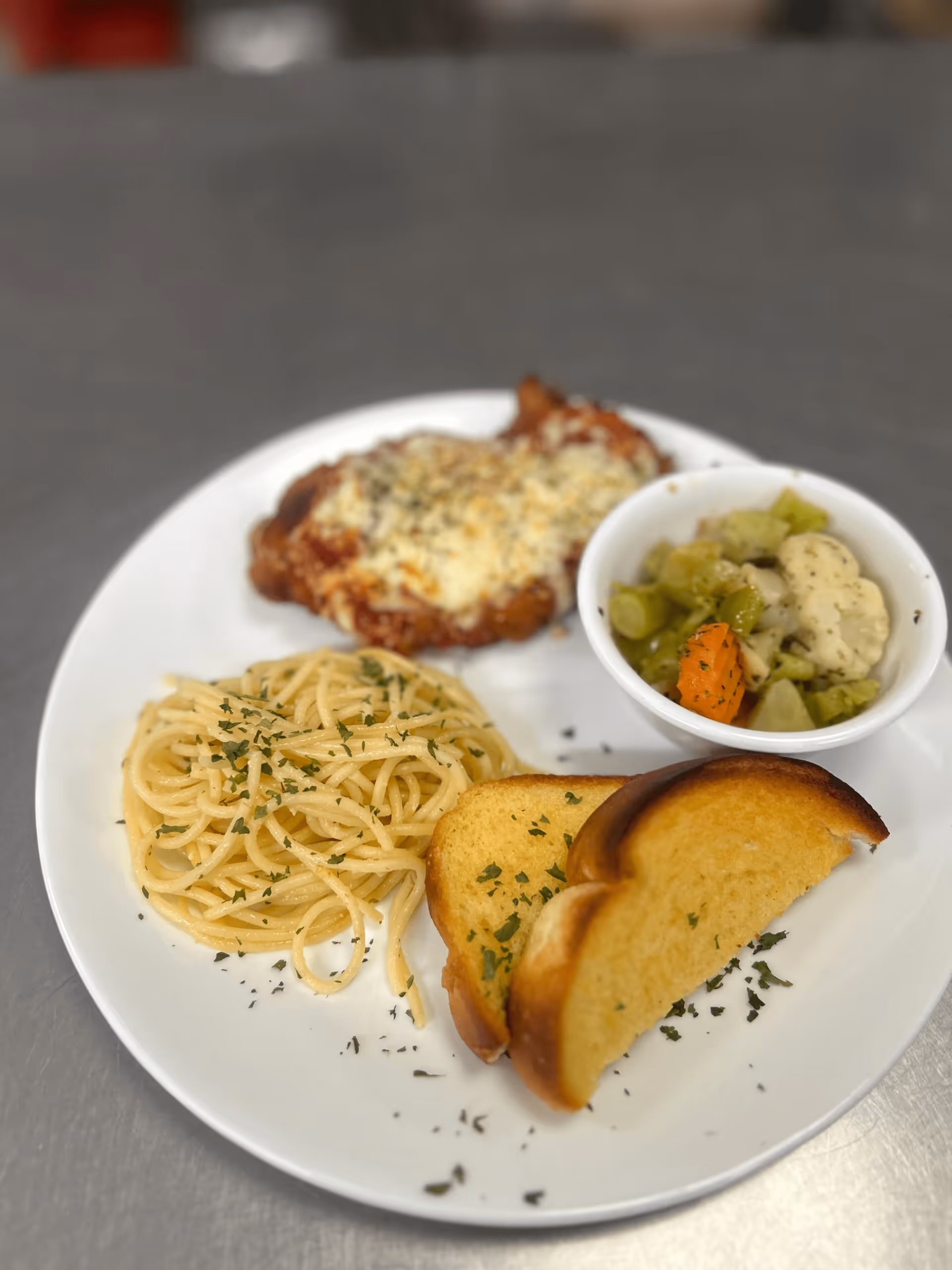 A white plate with a serving of spaghetti sprinkled with herbs, two slices of toasted garlic bread, a piece of chicken parmesan topped with melted cheese, and a small bowl of mixed steamed vegetables including carrots, cauliflower, and green beans.