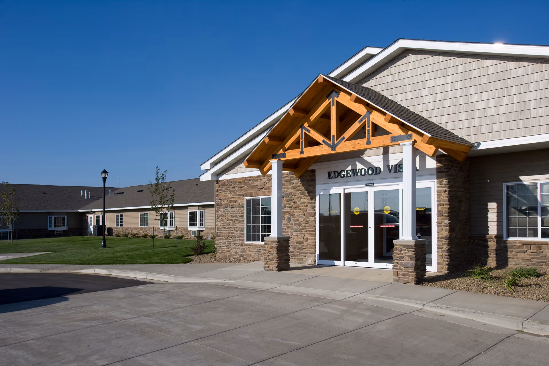 Exterior view of the entrance to Edgewood Bismarck Village, showing a building with stone and siding facade, a wooden gabled porch roof, glass double doors, and a paved driveway with green lawn and small trees under a clear blue sky.