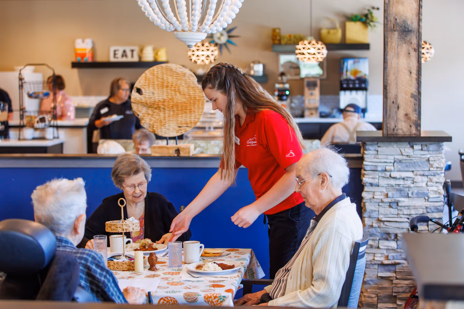 A caregiver in a red shirt serves food to elderly residents seated at a table in a dining area of a senior living facility. The table is covered with a patterned tablecloth and set with plates, cups, and glasses. The background shows other residents and a kitchen area with shelves and hanging lights.