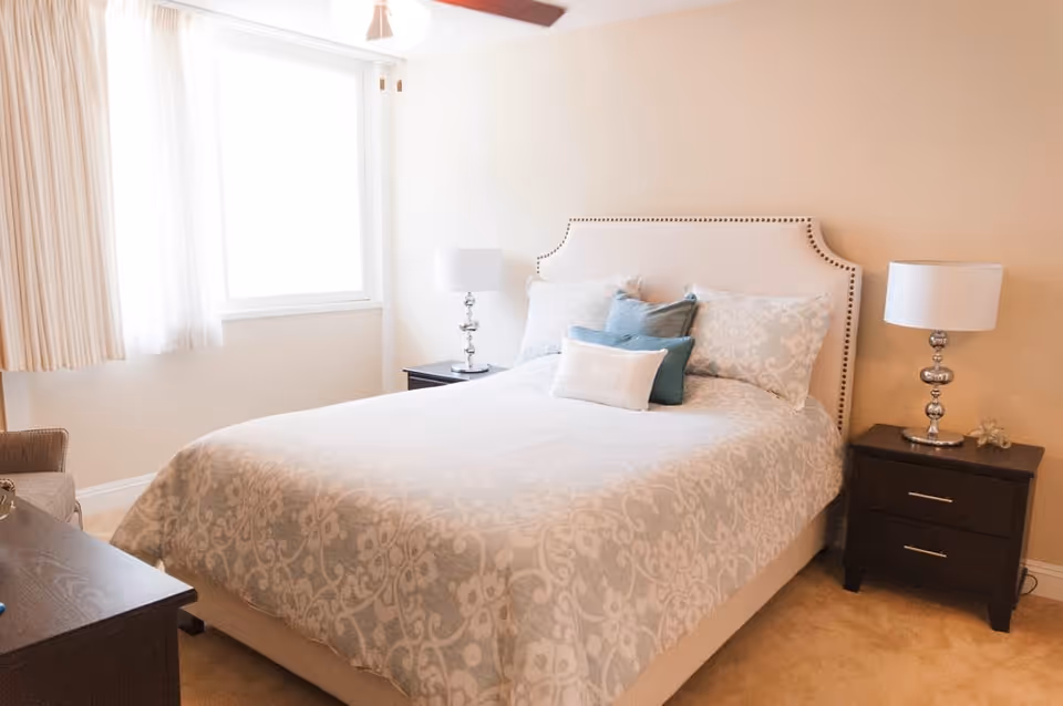 Bright, neutral bedroom featuring a bed with patterned bedding, decorative pillows, two nightstands with lamps, and a curtained window.