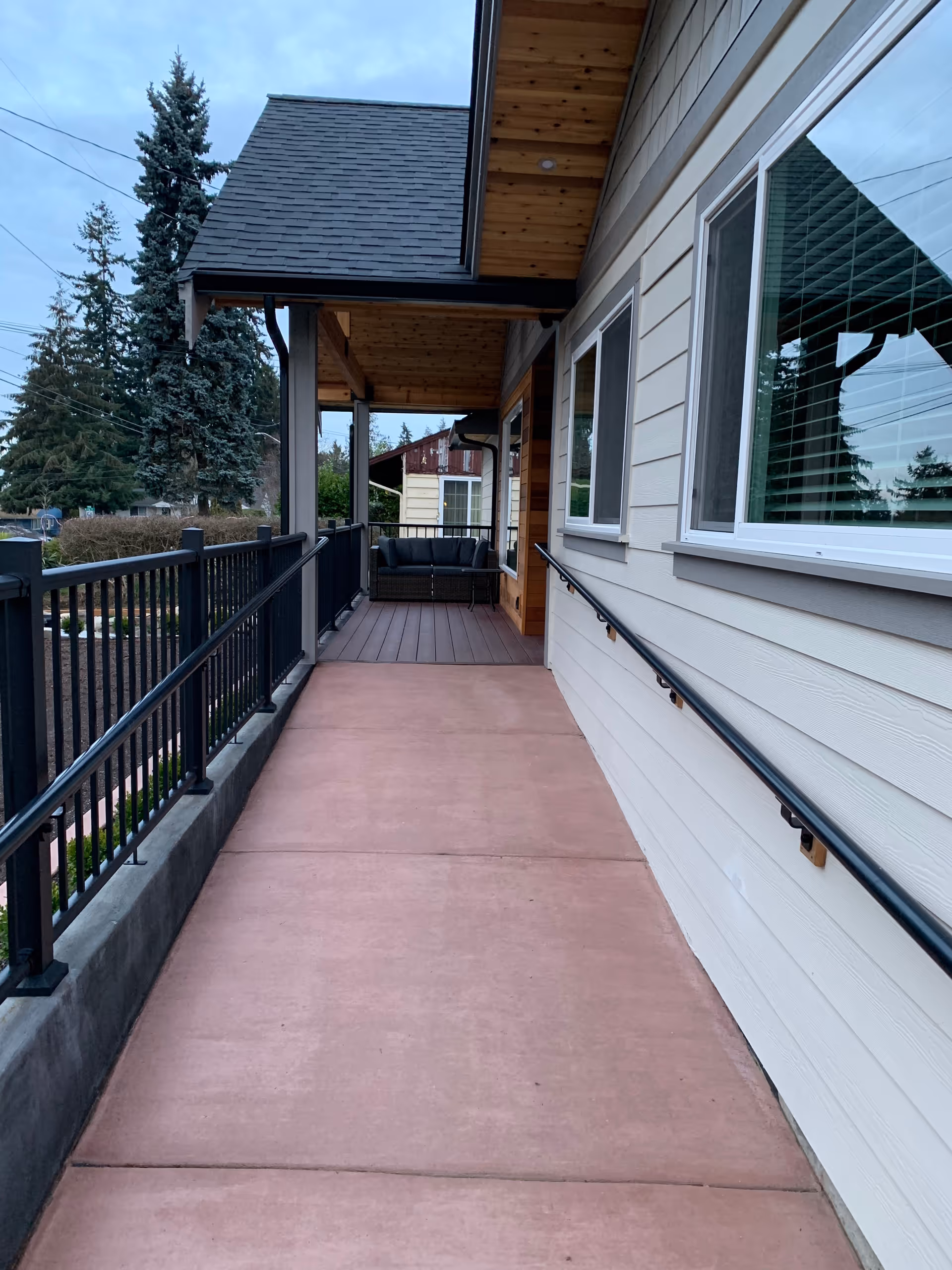 Covered outdoor walkway with a handrail on the right side attached to the building and a black metal railing on the left side. At the end of the walkway, there is a seating area with a black couch. The building has beige siding and white-framed windows. Trees and other buildings are visible in the background.