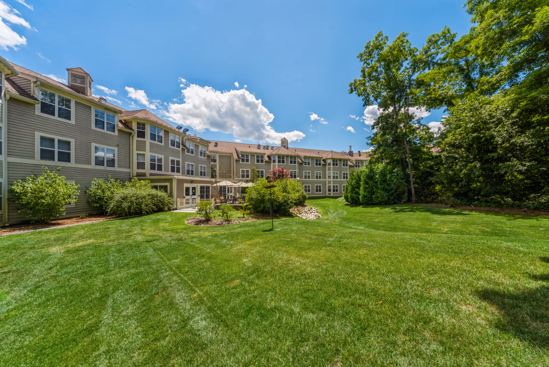 A large, well-maintained grassy lawn with bushes and trees surrounding a three-story residential building under a bright blue sky with some clouds.