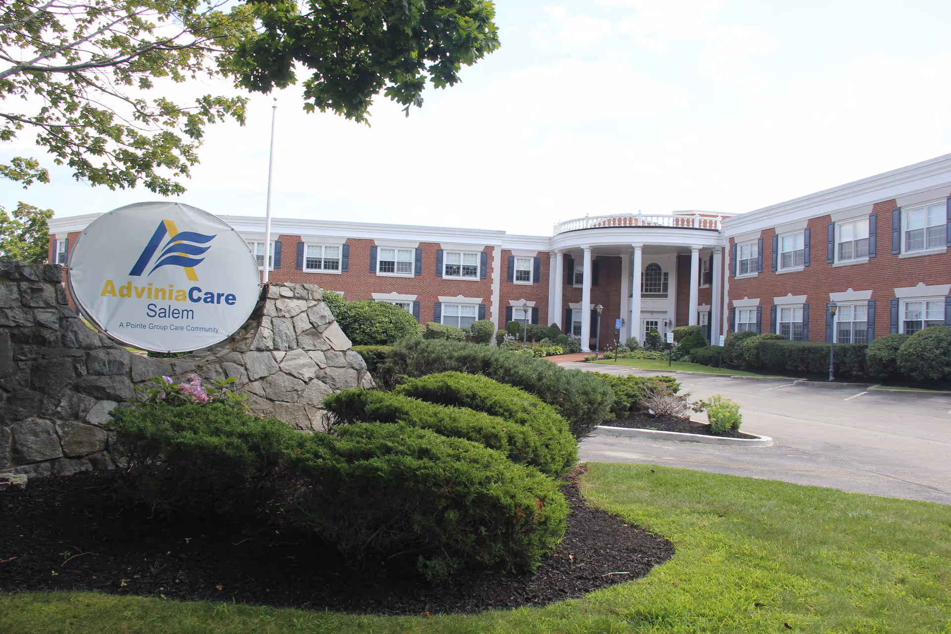 Front exterior view of the Adviniacare Salem facility, a large brick building with white trim and columns at the entrance, surrounded by well-maintained landscaping including bushes and trees. A stone sign with the Adviniacare Salem logo is visible in the foreground.