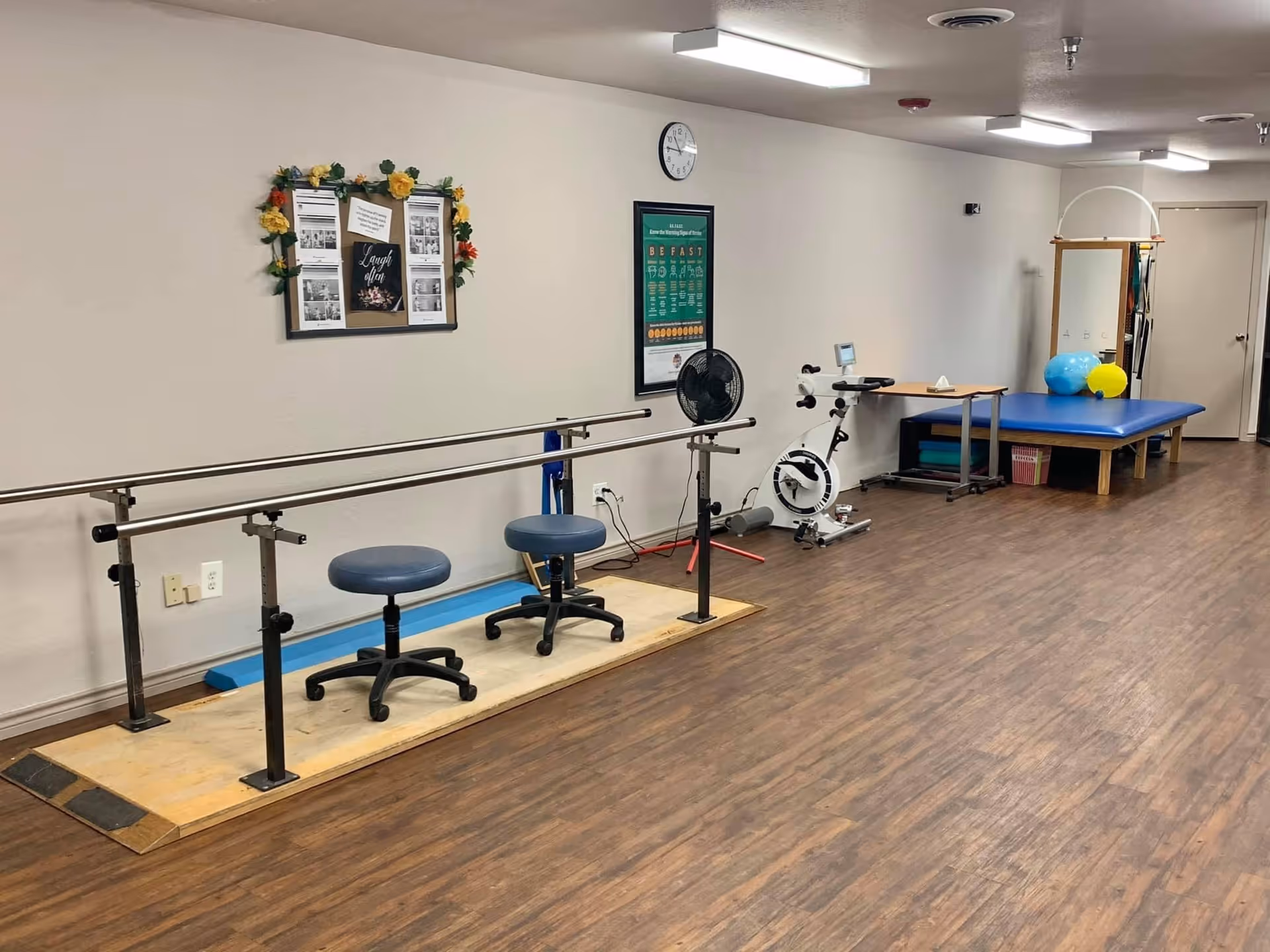 A physical therapy room with parallel bars for walking exercises, two blue stools, an exercise bike, a blue therapy table with exercise balls on it, a fan, and a bulletin board decorated with flowers on the wall.