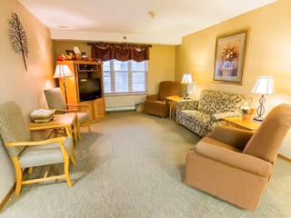 A cozy living room with beige walls and carpeted floor featuring a floral-patterned sofa, two armchairs, a wooden TV cabinet with a television, a side table with a lamp, and a window with brown curtains.