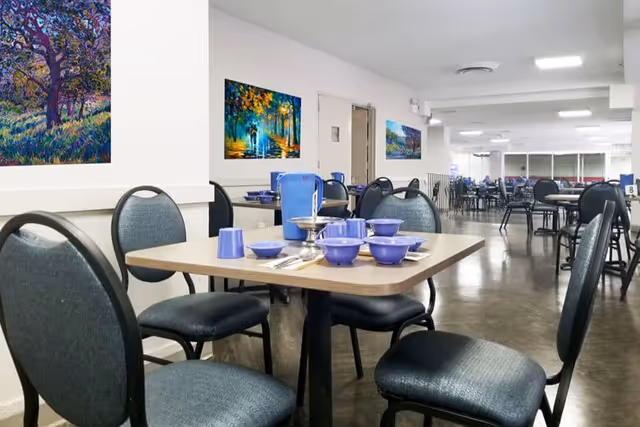 Interior view of a dining room in an assisted living facility with several tables and chairs. One table in the foreground is set with blue bowls, cups, and utensils. Colorful paintings hang on the white walls, and the room is well-lit with overhead lighting.