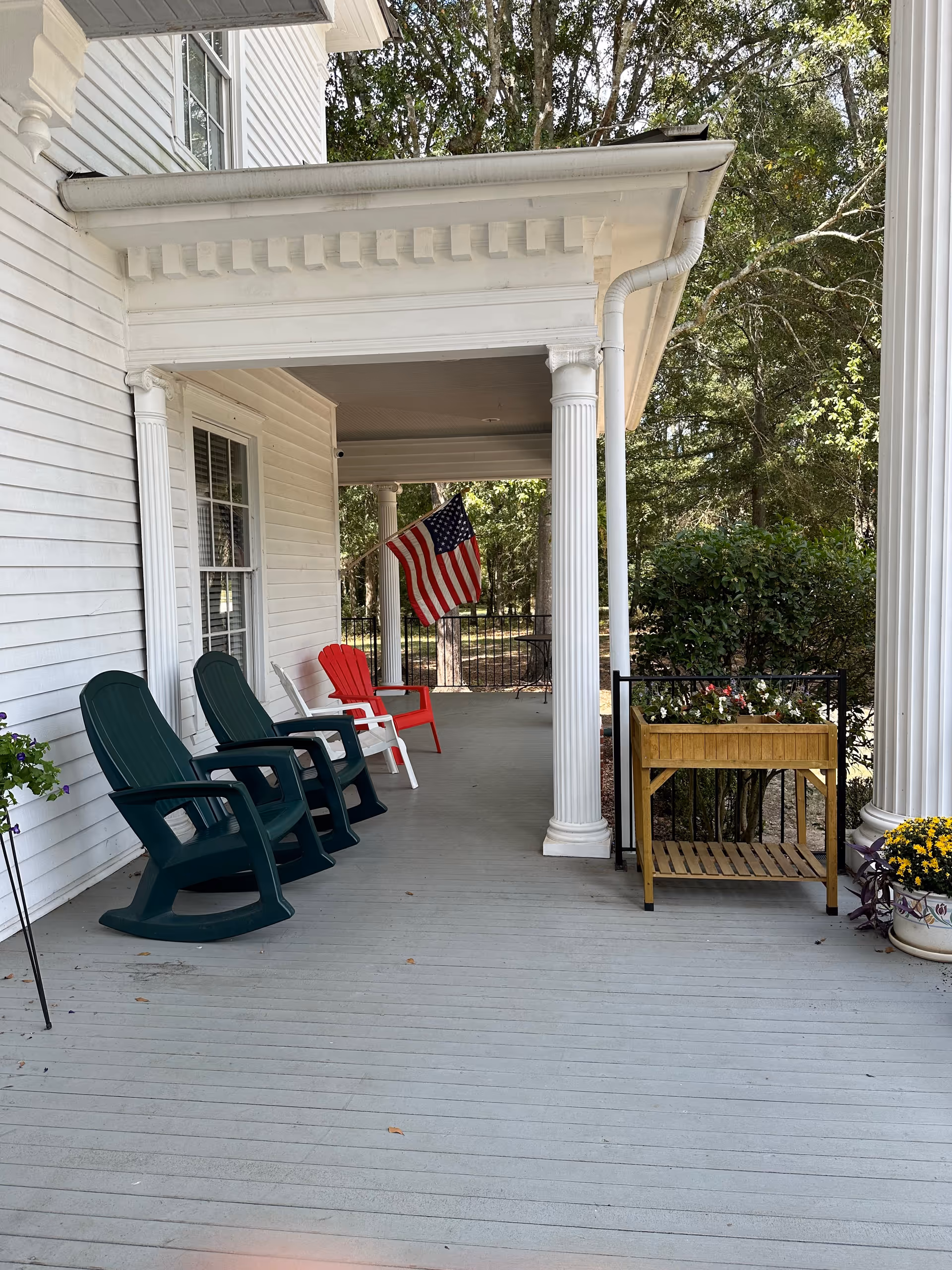 Covered front porch with white columns, several outdoor chairs, an American flag, and potted plants.