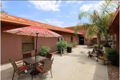 Sunlit courtyard patio with an umbrella-shaded table and chairs, benches, potted plants, and surrounding single-story buildings.