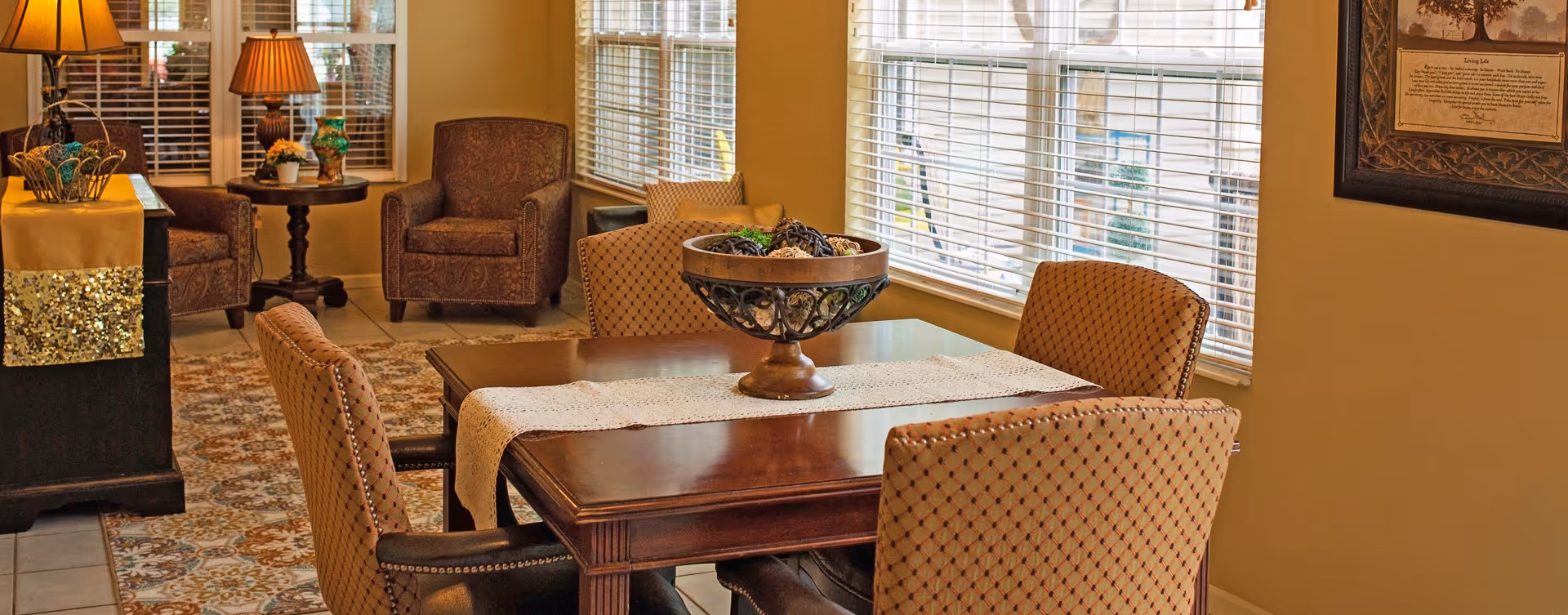 A cozy interior room with a wooden dining table surrounded by four upholstered chairs. The table has a decorative runner and a centerpiece bowl filled with decorative spheres. In the background, there are two patterned armchairs, a side table with a lamp and a vase, and large windows with blinds letting in natural light. The room has warm beige walls and a patterned area rug on the tiled floor.
