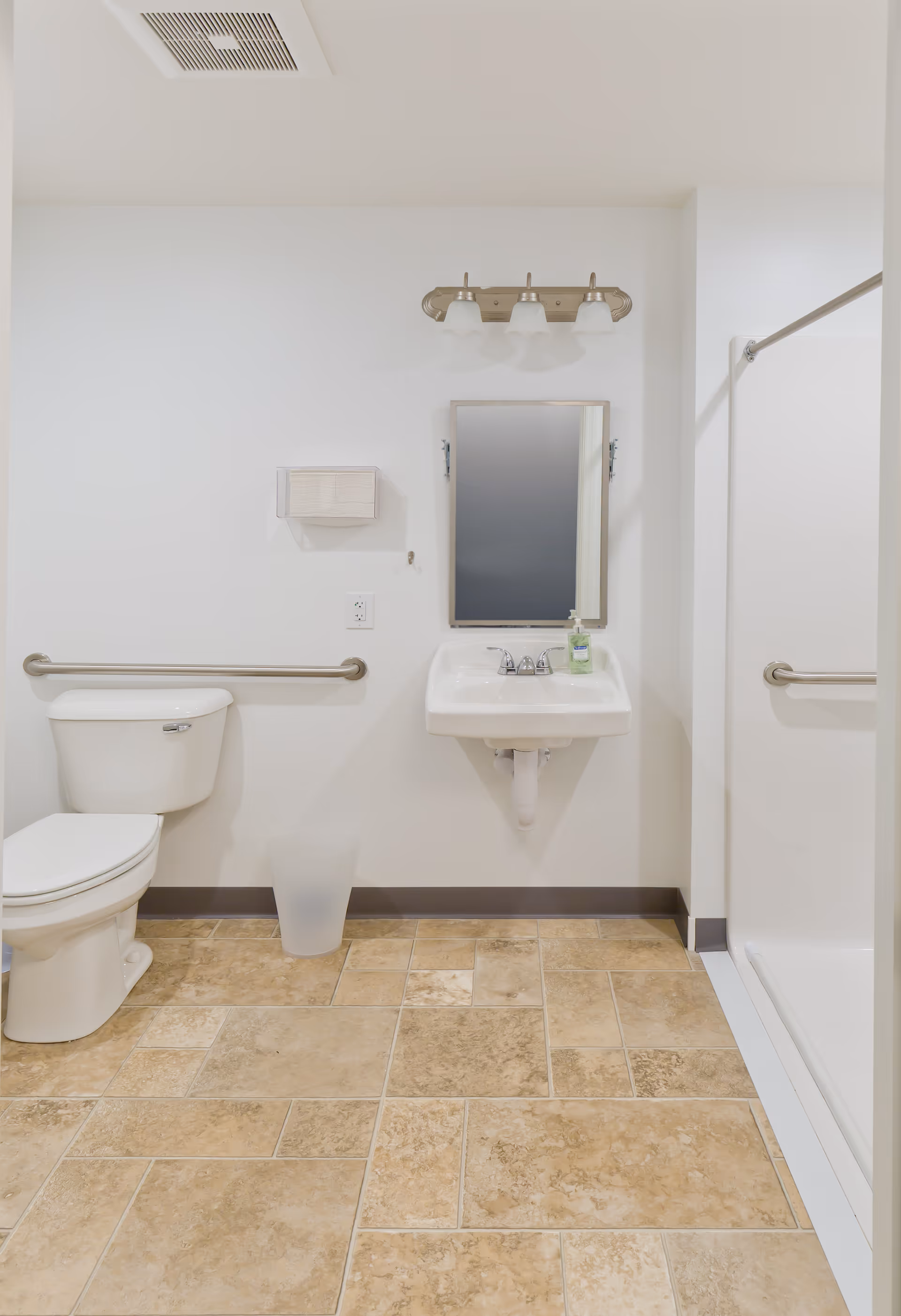 A clean and accessible bathroom with beige tiled floor, a white toilet with a horizontal grab bar on the wall beside it, a white wall-mounted sink with a mirror above it and a three-light fixture, a soap dispenser on the sink, and a walk-in shower with a grab bar on the right side.