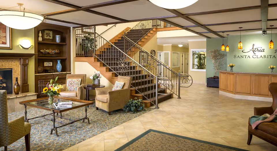 A spacious and well-lit senior living facility lobby featuring a seating area with armchairs and a glass coffee table with a flower vase. There is a decorative fireplace and built-in wooden shelves with vases and artwork. A staircase with ornate metal railings leads to an upper floor. The reception desk is visible on the right with pendant lights hanging above and a sign that reads 'Atria Santa Clarita' on the wall behind it.
