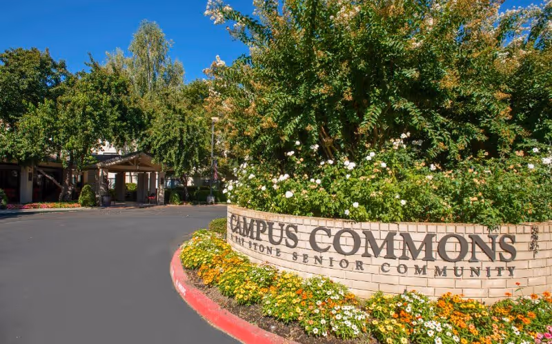 Entrance to Campus Commons senior living community with a brick sign surrounded by flowers and greenery, and a driveway leading to the building under a clear blue sky.