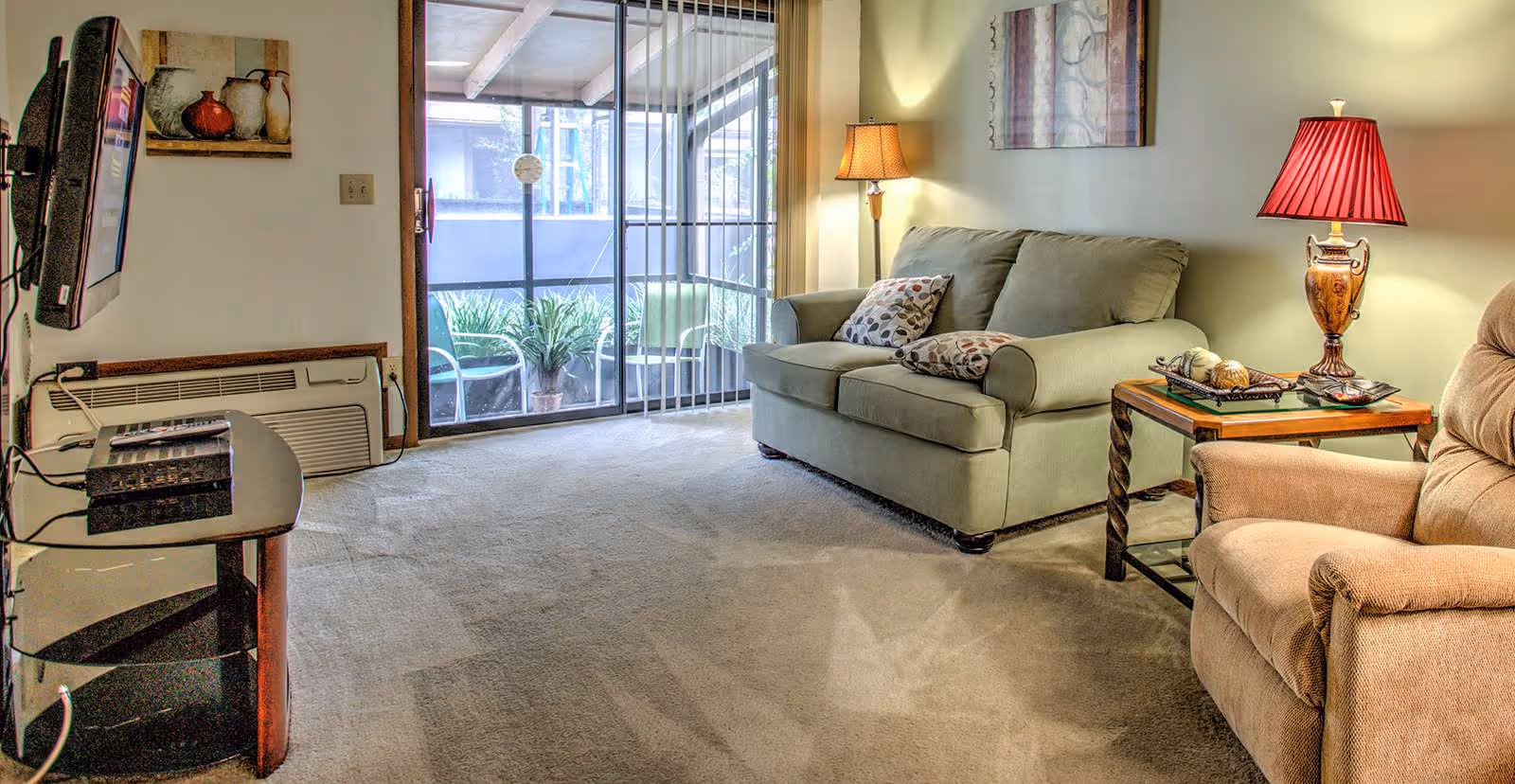 A cozy living room with a green sofa adorned with patterned pillows, a beige recliner chair, and a wooden side table with a decorative lamp and books. There is a floor lamp next to the sofa, a wall-mounted TV on the left, and a sliding glass door leading to a small balcony with plants outside.