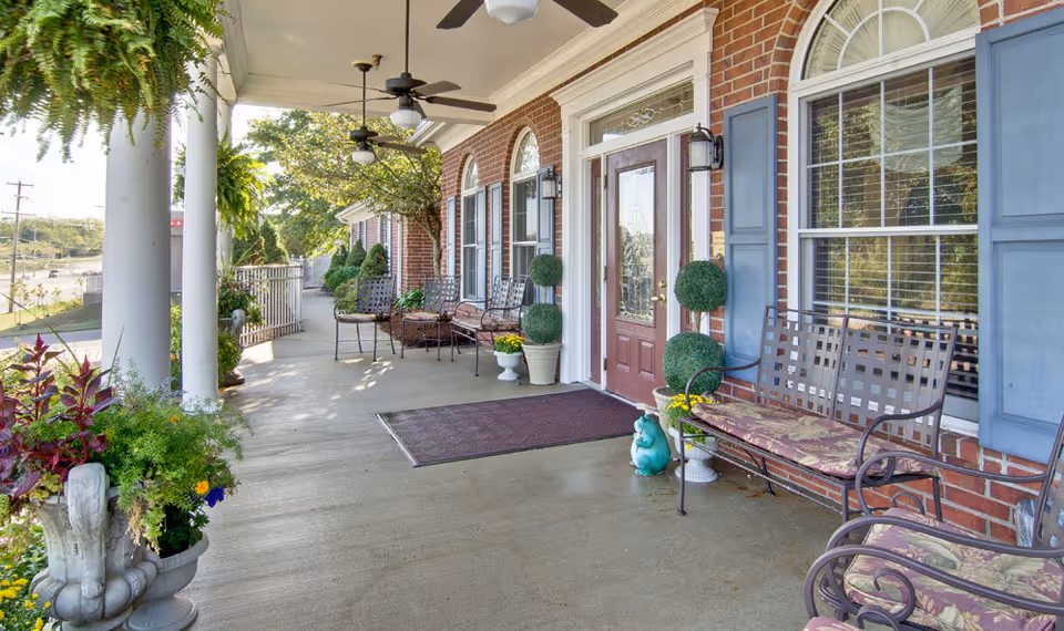 A spacious covered porch at Ravenwood Senior Living with brick walls, blue shutters, and a glass-paneled front door. The porch features several cushioned metal benches, potted plants, hanging ferns, ceiling fans, and outdoor lighting fixtures, creating a welcoming and comfortable outdoor seating area.