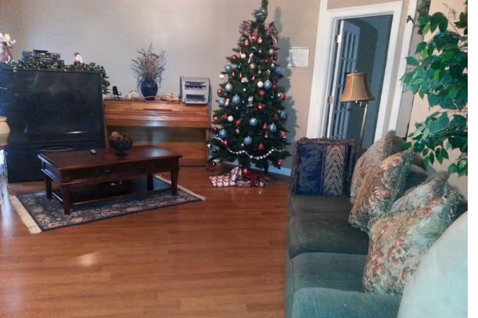 A cozy living room decorated for Christmas with a decorated Christmas tree, a green sofa with floral cushions, a wooden coffee table on a small rug, a piano against the wall, and a floor lamp near a doorway.