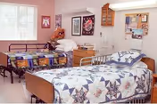 A shared bedroom in a senior care facility with two hospital-style beds, each covered with a colorful quilt. The room has a window with vertical blinds, a wooden nightstand between the beds, and various decorations on the walls including framed pictures and a bulletin board.