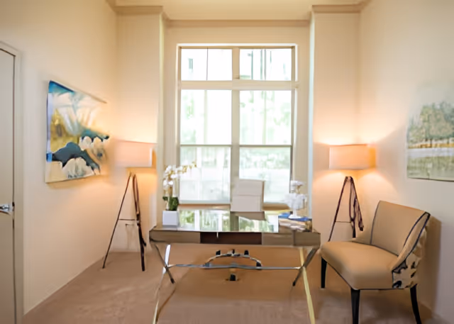 A bright office or study room with a large window letting in natural light. The room features a glass desk with a white chair behind it, two tripod floor lamps with beige lampshades on either side, a beige upholstered chair to the right, and two abstract paintings on the walls.
