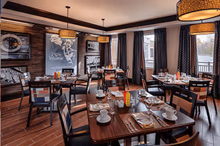 Dining room with several wooden tables and chairs set with white cups, plates, and silverware. The room has large windows with dark curtains, pendant lights hanging from the ceiling, and framed artwork on the walls.