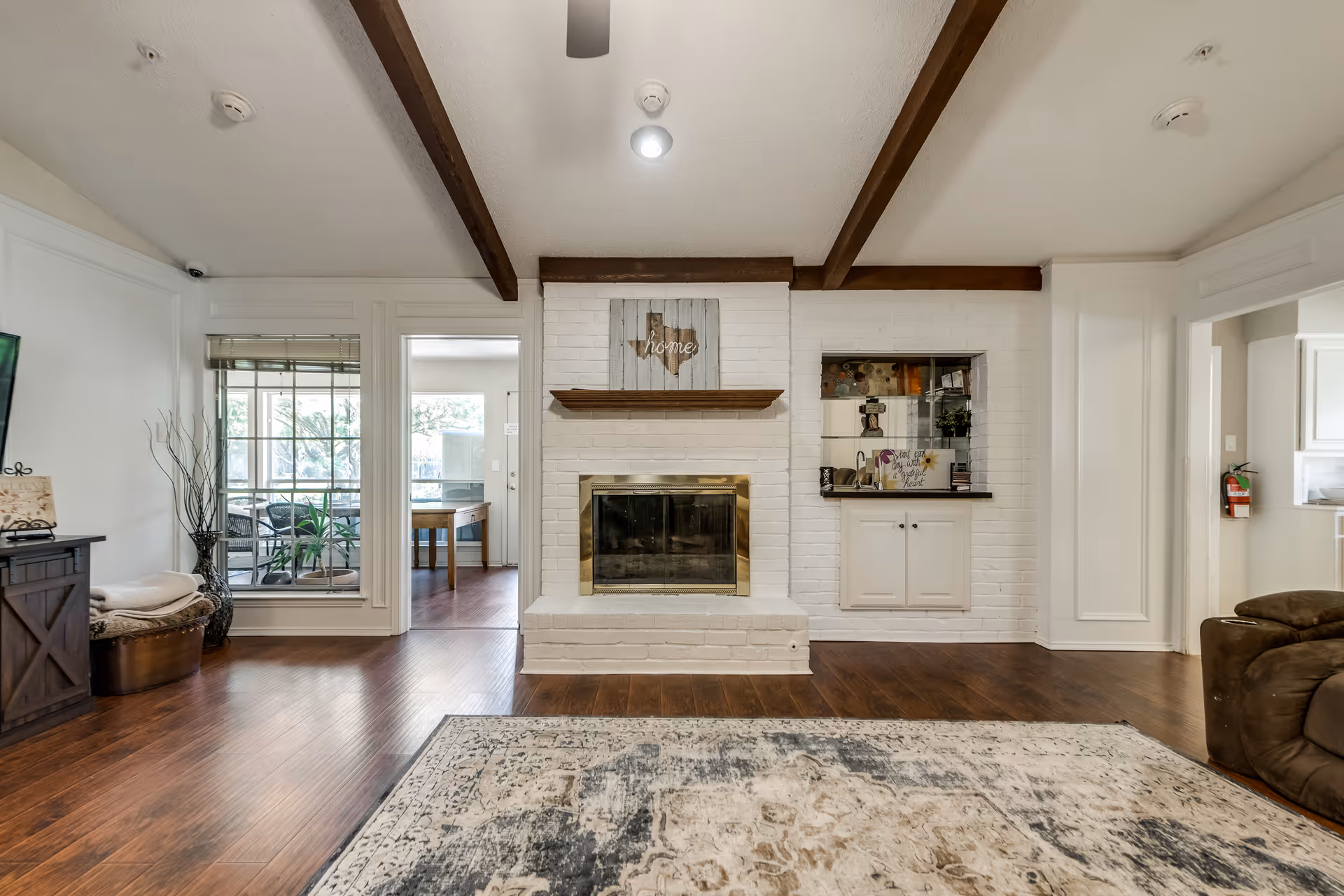A cozy living room with wooden beams on the ceiling, a white brick fireplace with a wooden mantel, and a decorative sign above it. To the right of the fireplace is a built-in cabinet with shelves displaying decor items. The room has wooden flooring and a patterned area rug in front of the fireplace. There are large windows and doorways leading to other rooms, with some furniture partially visible.