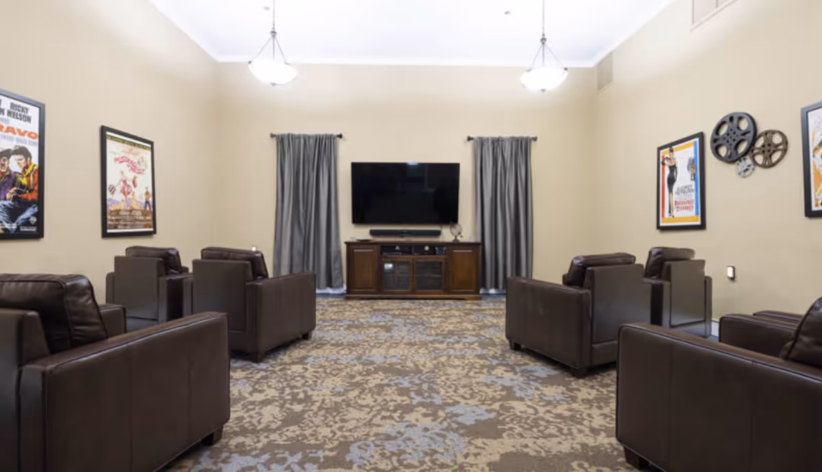 Community media room with rows of brown leather armchairs facing a wall-mounted TV, framed movie posters, and patterned carpet.