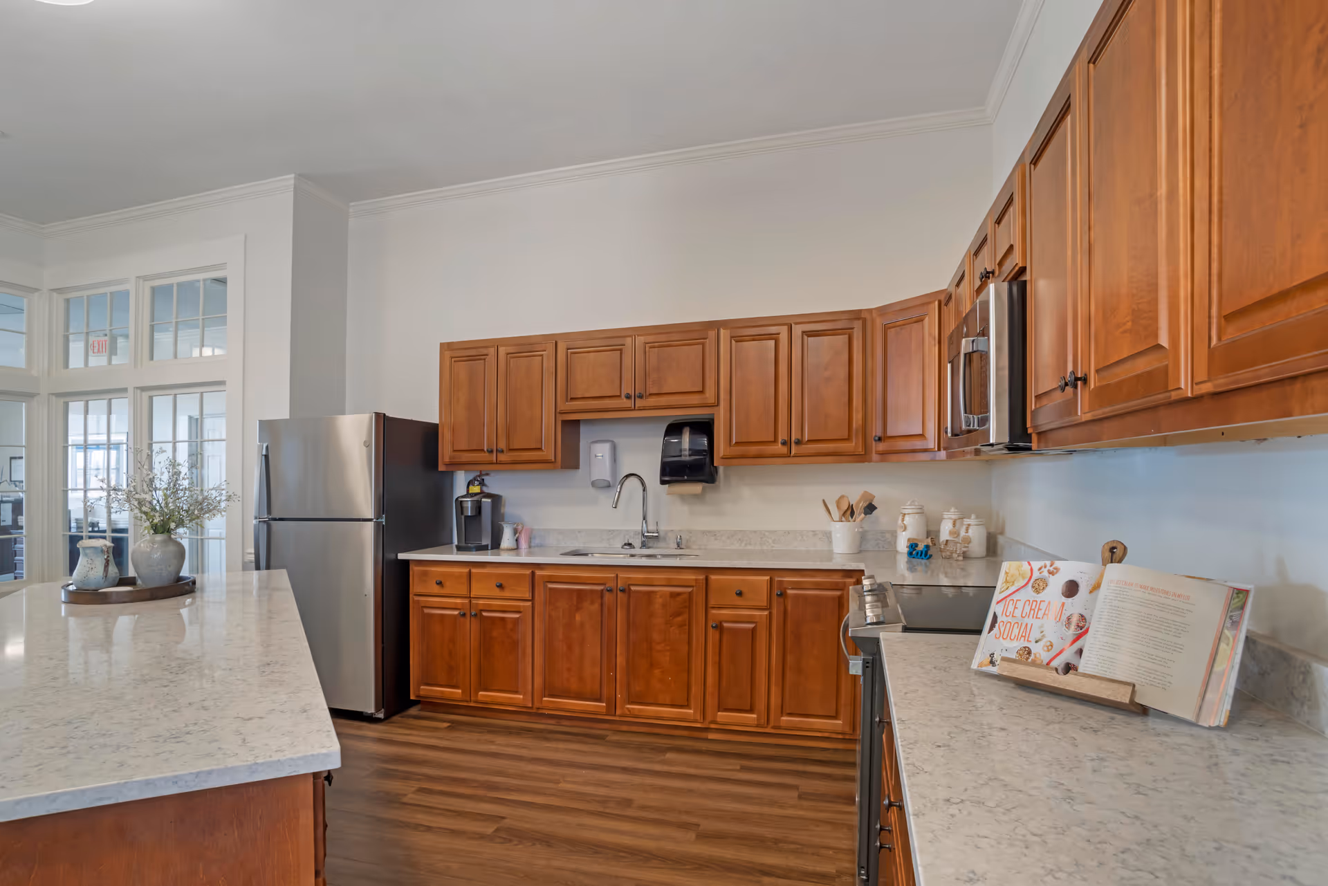A spacious kitchen with wooden cabinets, a stainless steel refrigerator, a microwave, and a stove. The countertops are light-colored with a marble-like pattern. There is a kitchen island with decorative vases and a tray. A cookbook is open on a stand on the counter. The floor is wooden, and there are glass-paneled doors in the background.