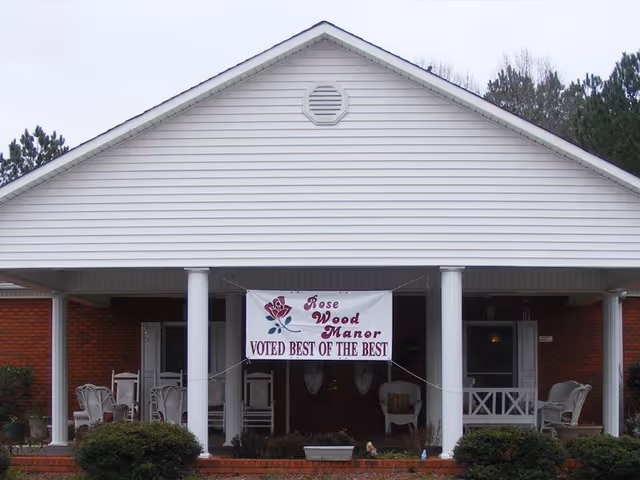 Front exterior view of a single-story building with white siding and brick walls, featuring a covered porch supported by white columns. A banner hanging between the columns reads 'Rose Wood Manor Voted Best of the Best'. There are chairs and small tables on the porch, and some bushes in front of the building.