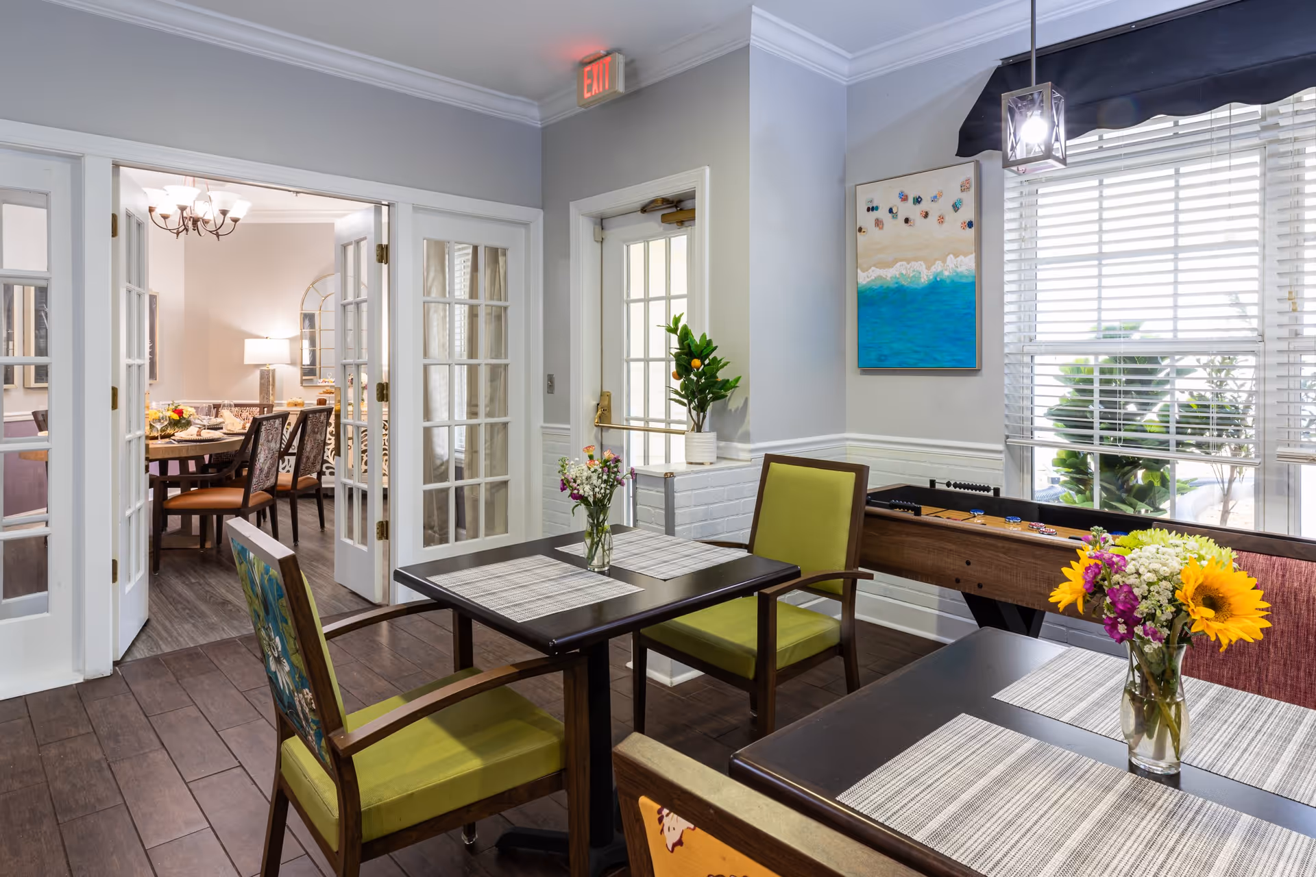 A bright and inviting interior common area with two tables and chairs, each table adorned with a small vase of flowers. There is a foosball table against the wall beneath a window with white blinds. A colorful painting of a beach scene hangs on the wall. Through open French doors, a dining room with a chandelier and a set dining table is visible.