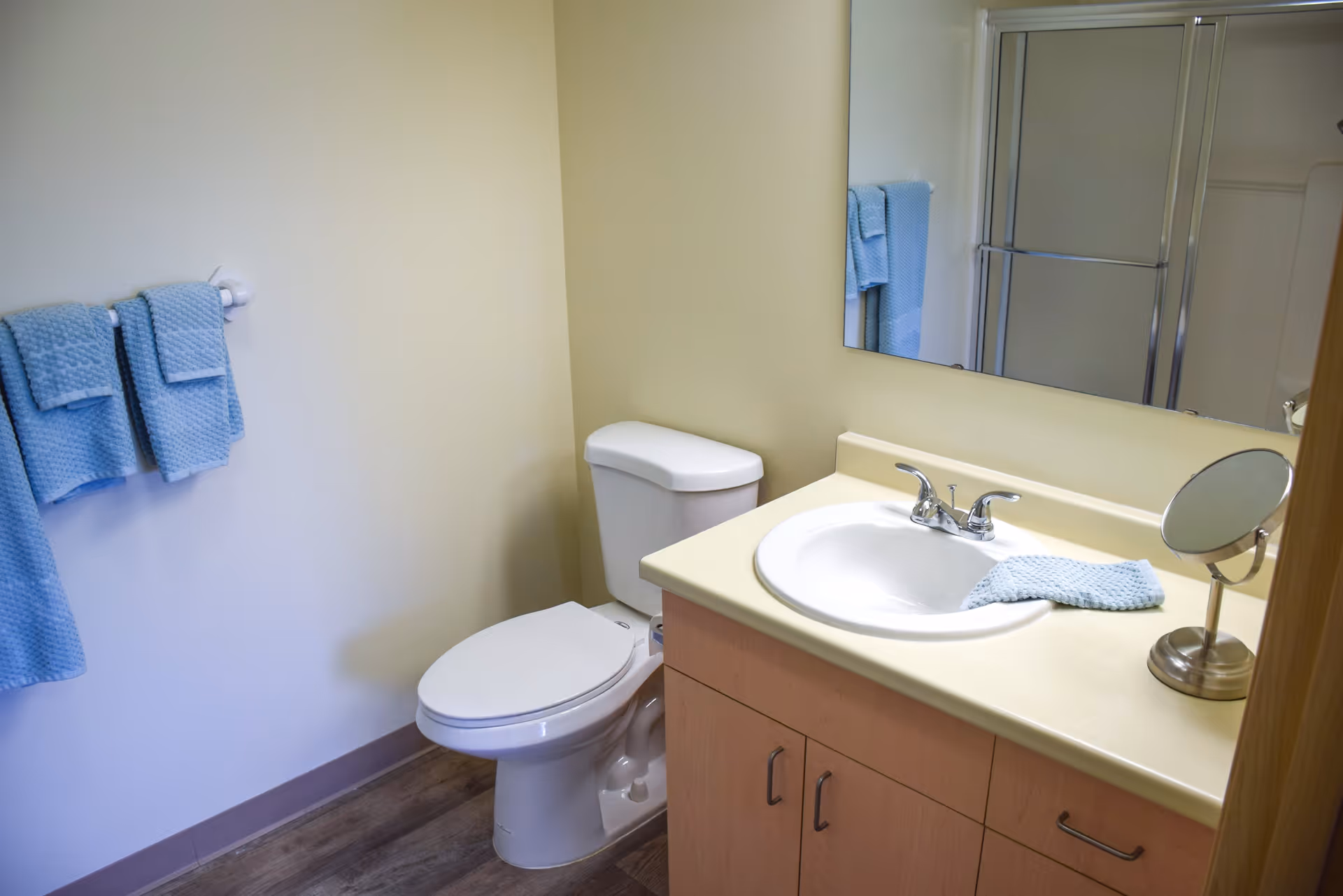 A clean bathroom with a white toilet, a sink with a beige countertop, a small round mirror, and blue towels hanging on a towel rack and placed on the sink. There is a glass shower door visible in the mirror reflection.