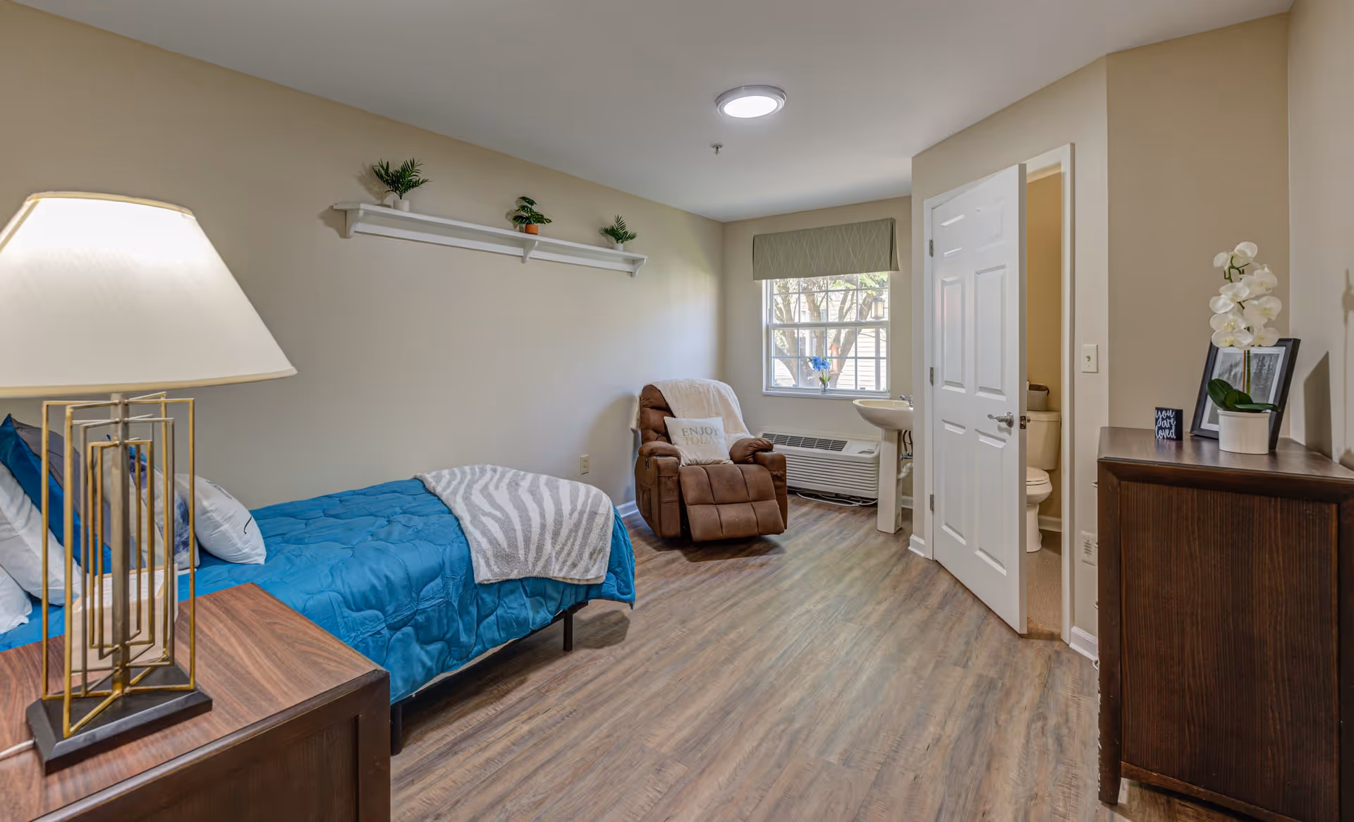 A cozy bedroom in a senior living facility featuring a single bed with blue bedding and pillows, a wooden nightstand with a lamp, a comfortable brown recliner chair with a white throw blanket and pillow, a window with a green valance, a small sink, and an open door leading to a bathroom. The room has wood flooring, beige walls, and decorative plants on a wall shelf and dresser.