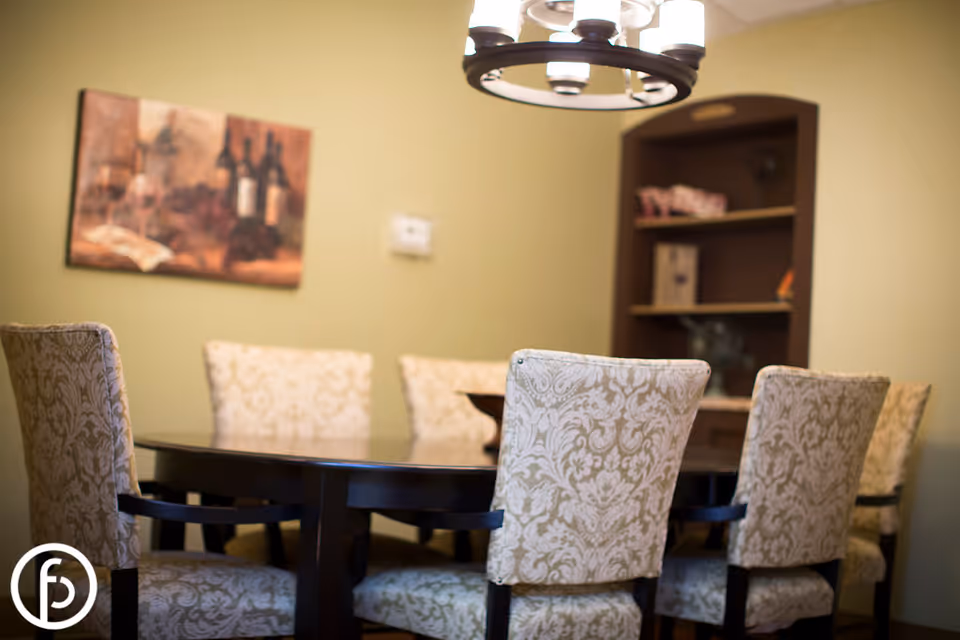 A dining room with a dark wooden oval table surrounded by six upholstered chairs with a beige floral pattern. A chandelier with multiple lights hangs above the table. In the background, there is a wooden shelving unit with decorative items and a framed painting of wine bottles and glasses on the wall.