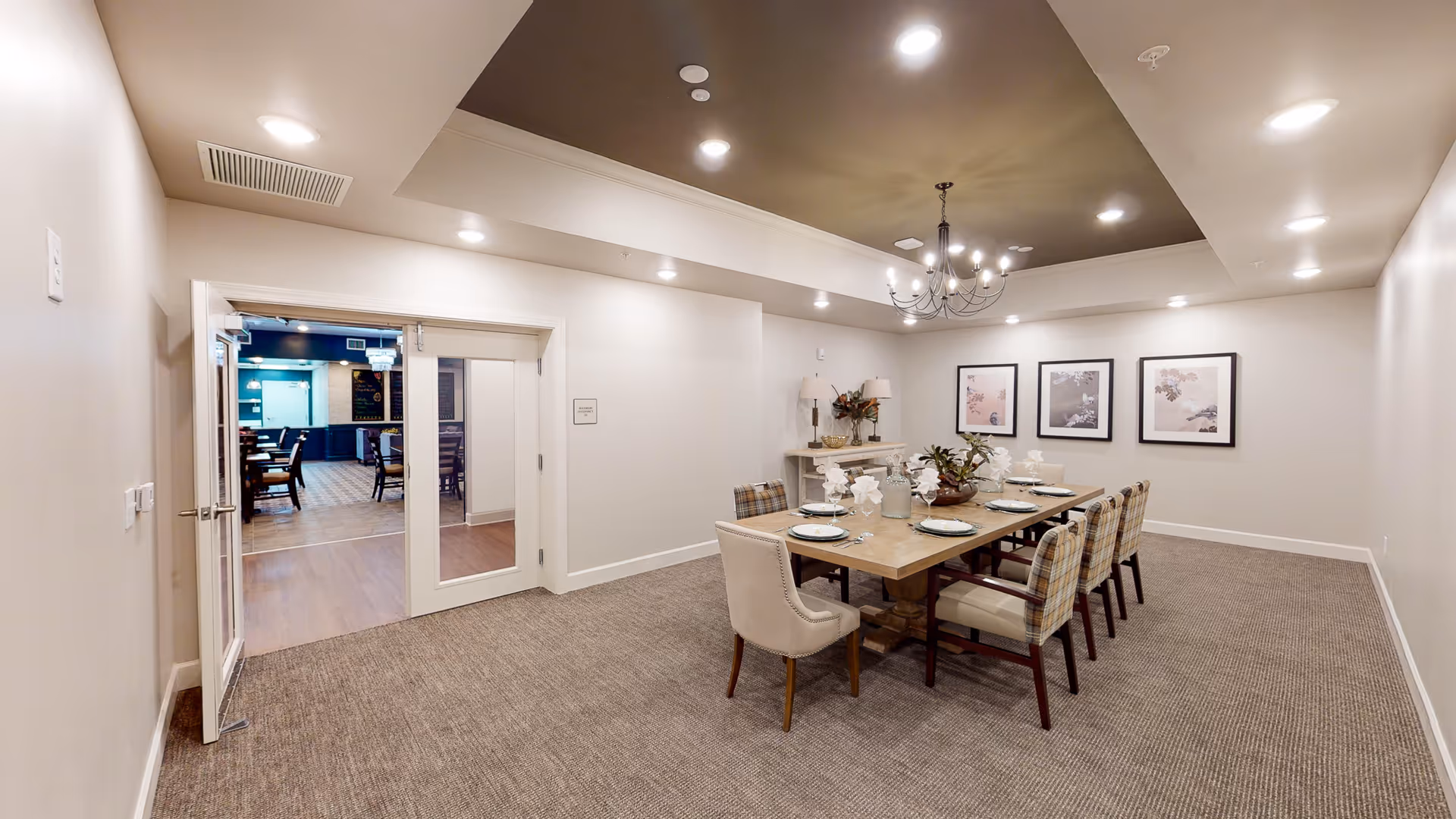 Well-lit private dining room with a long table set for guests, upholstered chairs, a chandelier, and double doors leading to an adjacent room.