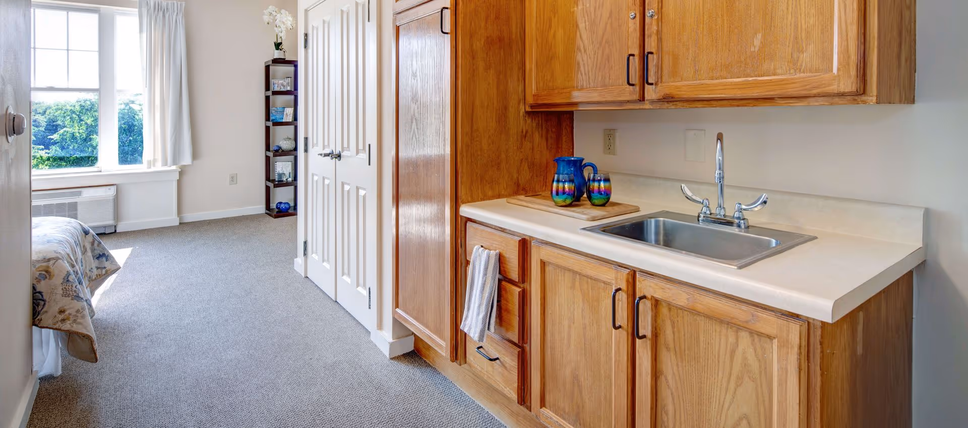 Interior view of a senior living facility room showing a small kitchenette with wooden cabinets, a sink, and a countertop with a colorful pitcher and glass. To the left, there is a carpeted area leading to a window with white curtains and a bed partially visible.