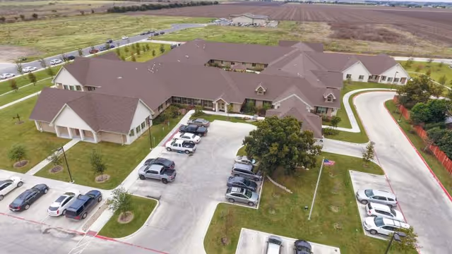 Aerial view of Legend Oaks Healthcare And Rehabilitation - New Braunfels showing a large single-story building with a brown roof surrounded by parking lots and green lawns. Several cars are parked in the parking areas, and there is a flagpole with an American flag near the entrance. The surrounding area includes open fields and roads.