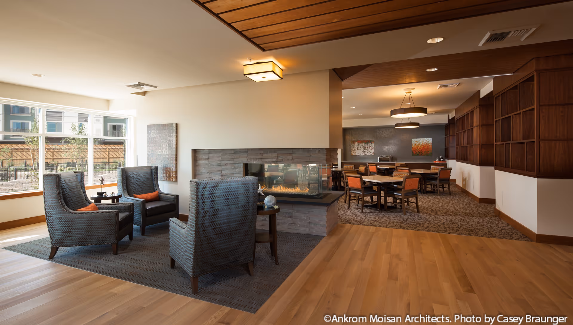 A cozy senior living facility common area with four gray armchairs arranged around a small table near a modern glass-enclosed fireplace. Large windows let in natural light, and in the background, there is a dining area with several tables and chairs, warm lighting, and wooden shelving on the walls.