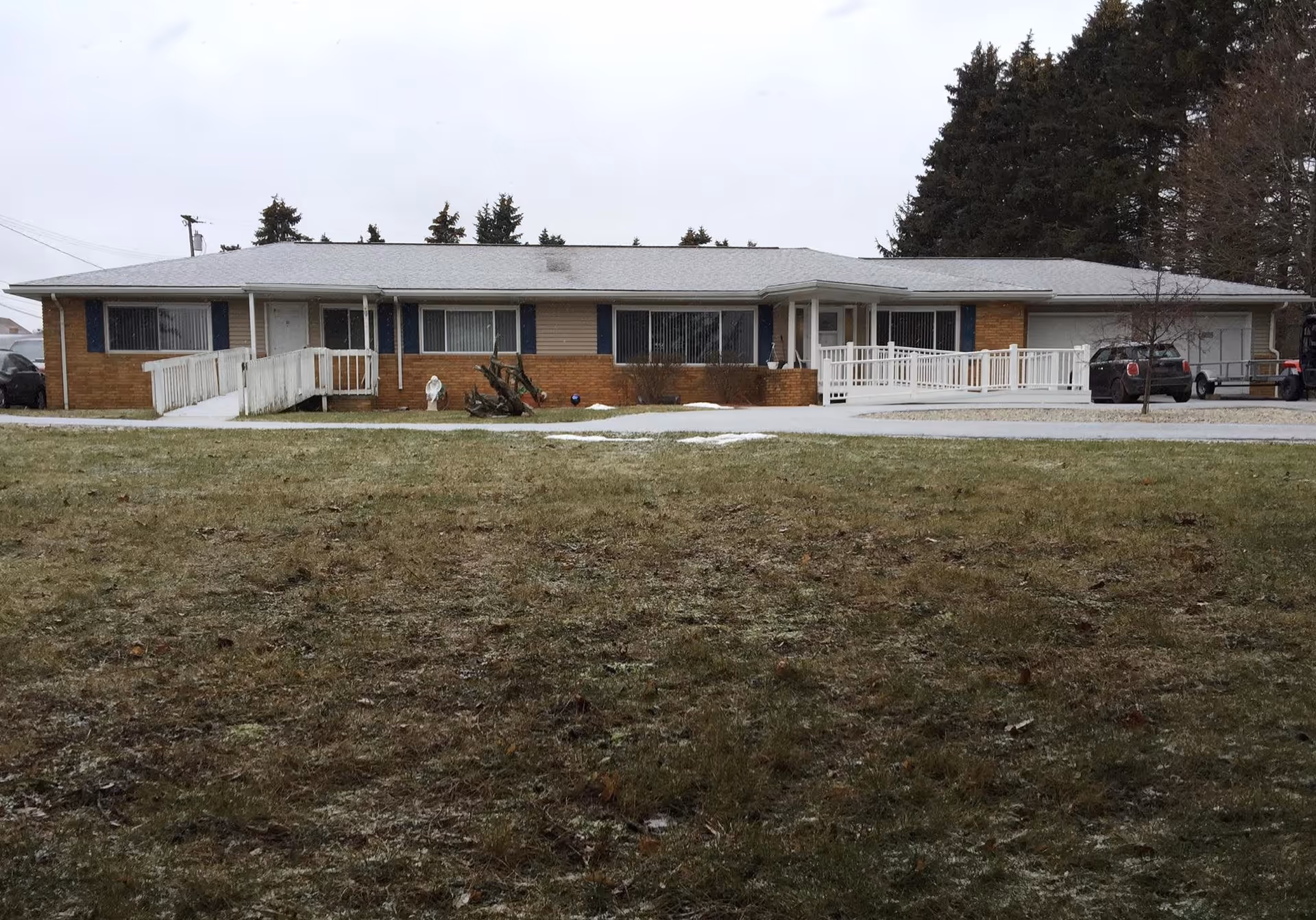 Single-story brick building with a gray roof and multiple windows, featuring two white wheelchair ramps leading to separate entrances. There is a grassy area in the foreground and several tall evergreen trees in the background. A few vehicles are parked near the building.